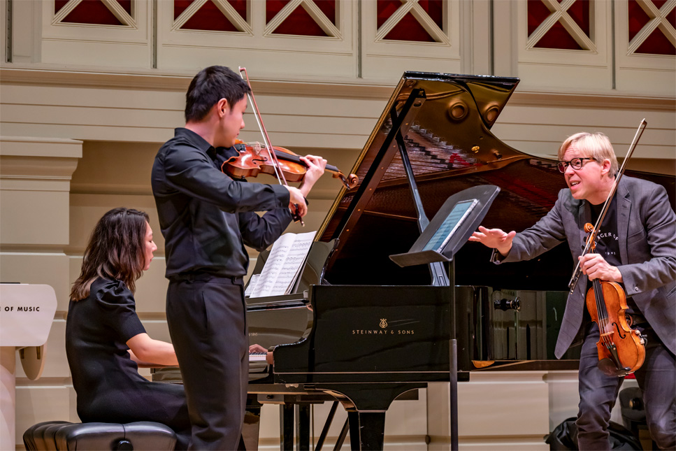 A male violinist and a female pianist performing on stage, with a male violin teacher teaching them to their right, on a well-lit stage.